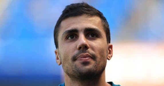 رودري لاعب مانشستر سيتي بقميص إسبانيا، وخلفه شعاري برشلونة وريال مدريد